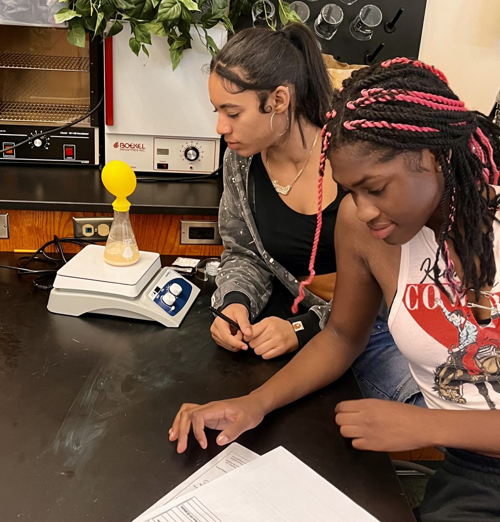 Two students watch balloons inflate over a flask